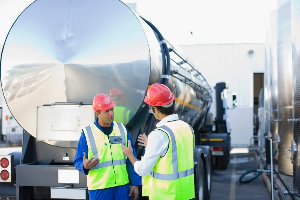 workers-talking-by-tanker-truck-2025-04-04-08-08-35-utc 1 - Rhino Fuel Two fuel delivery workers in safety gear discussing how fuel delivery works beside a large fuel tanker truck at an industrial facility