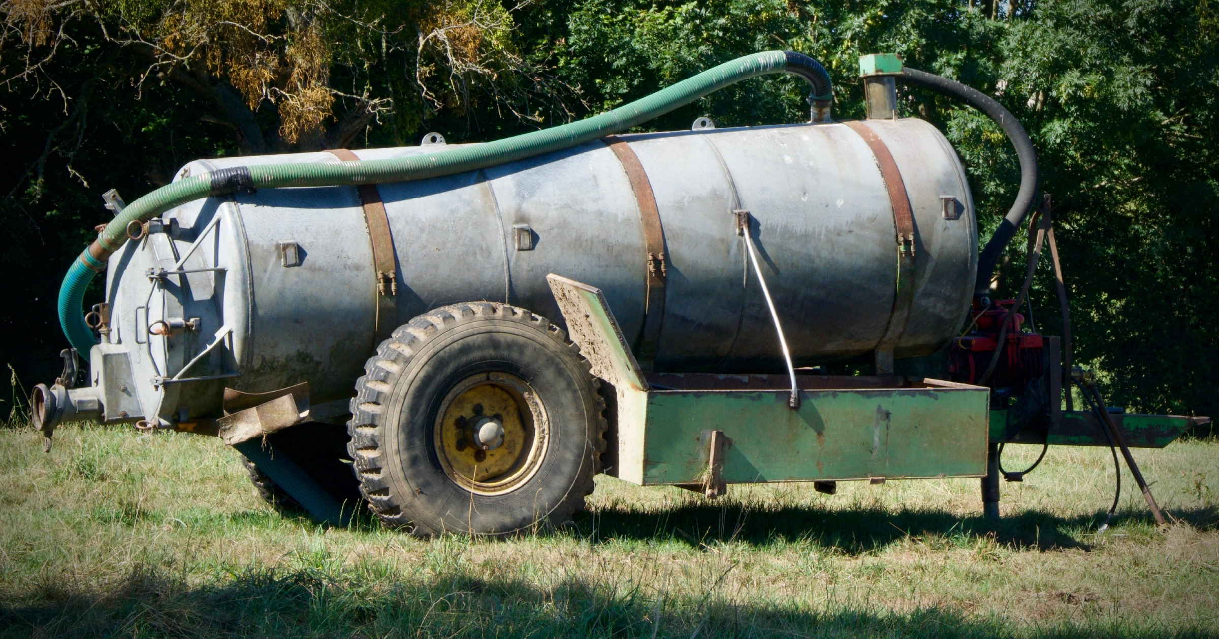 Farm fuel tanker trailer with hoses parked in a grassy field near trees.