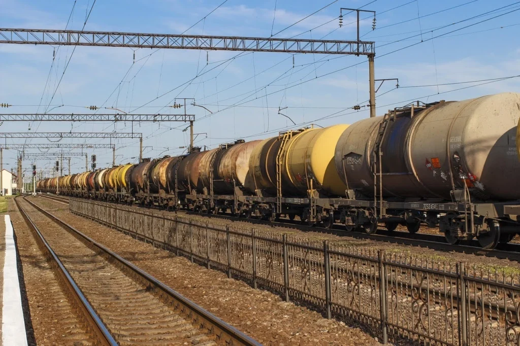 A long line of yellow and gray tanker rail cars stretches along the tracks in a rail yard beneath overhead electrical lines.